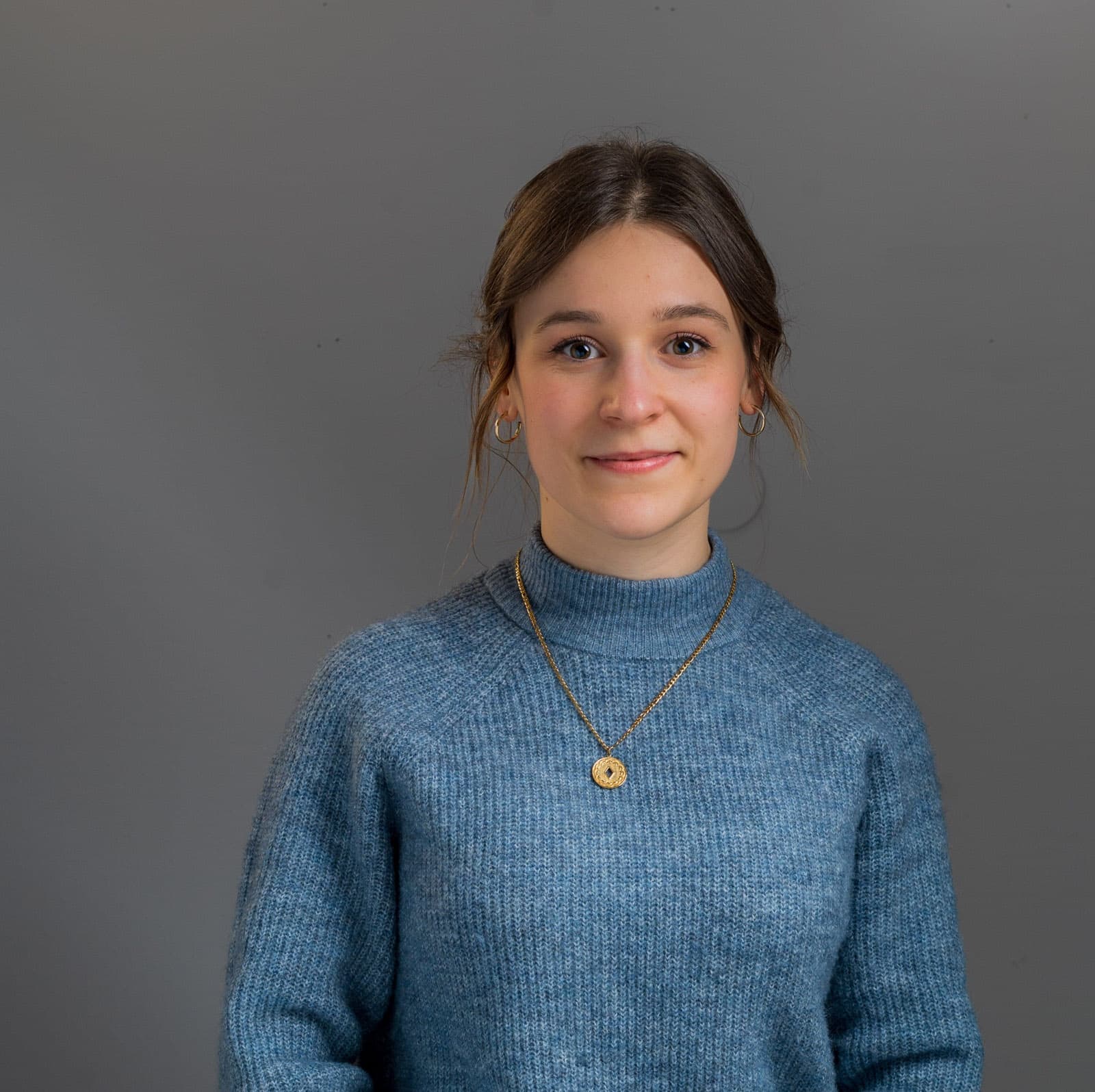 Junge Frau mit blauem Strickpullover und goldener Münz-Halskette vor grauem Studiohintergrund