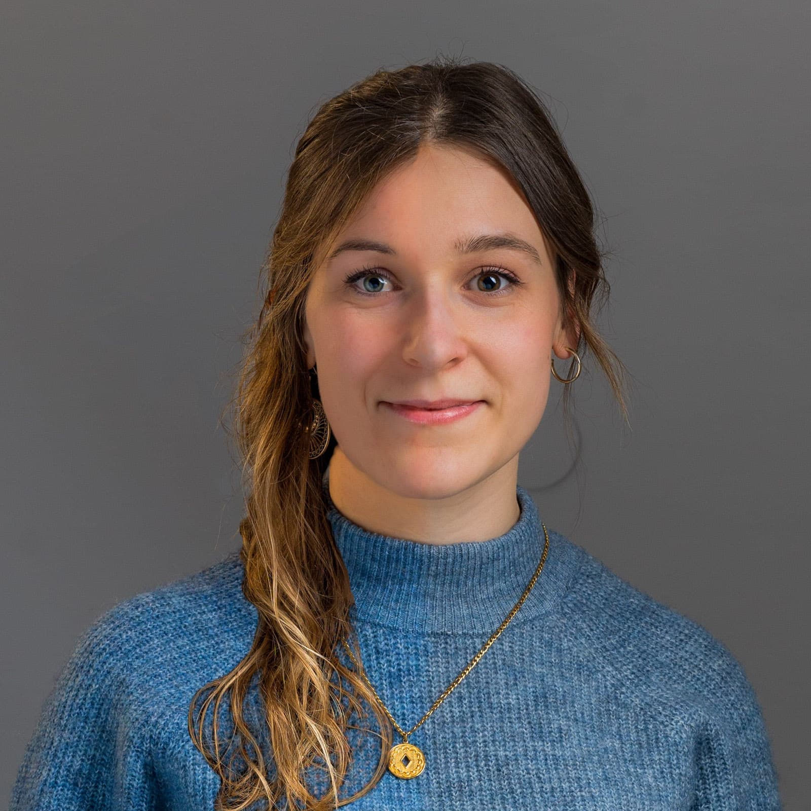 Lächelnde Frau mit Zopf und blauem Strickpullover vor grauem Studiohintergrund