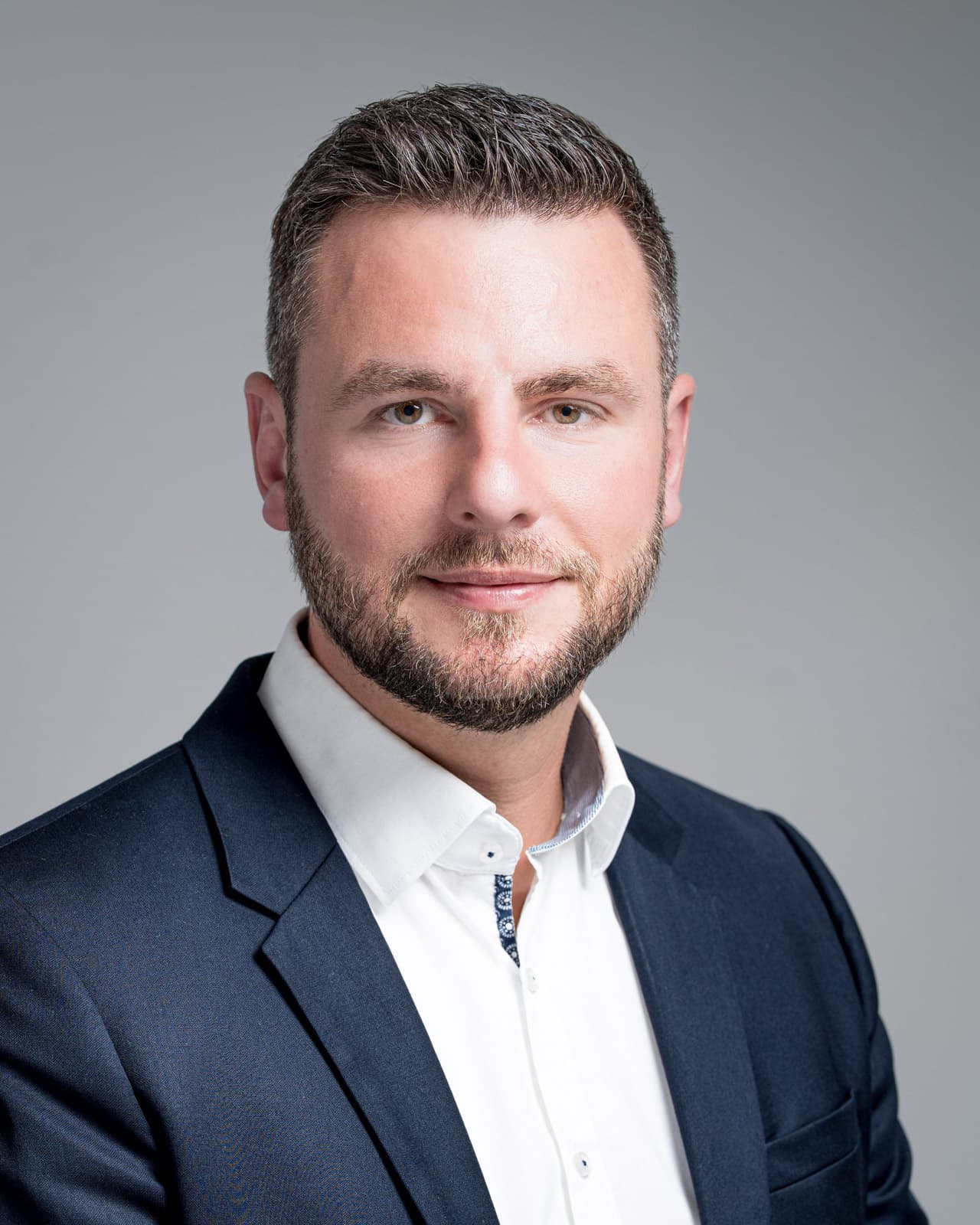 Geschäftsmann mit Vollbart, dunklem Sakko und weißem Hemd, freundliches Headshot-Lächeln