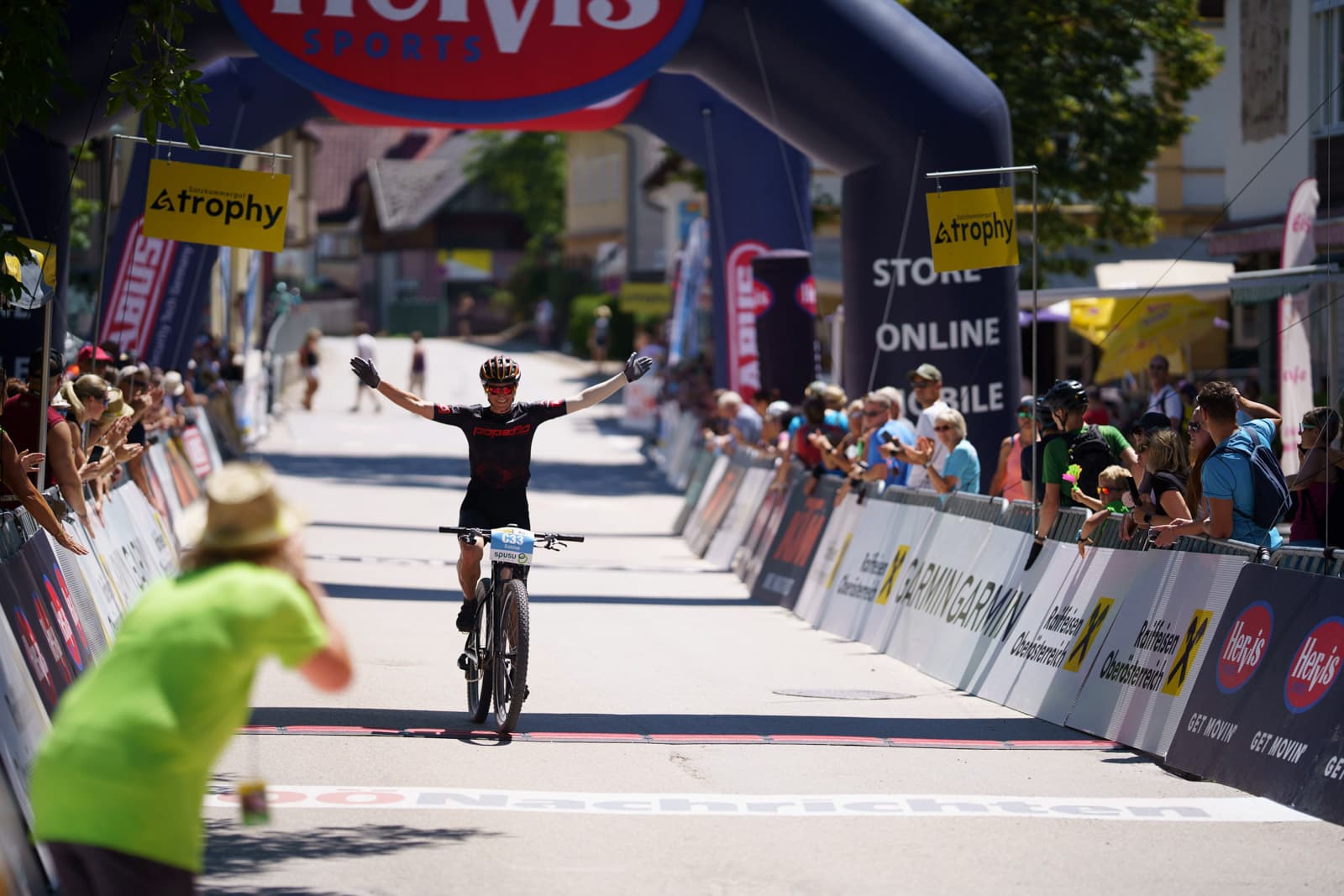 Mountainbiker fährt mit erhobenen Armen unter Hervis-Sports-Zielbogen ins Ziel