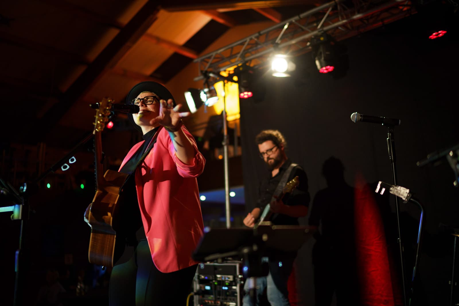 Sängerin mit Hut, Brille und pinkem Blazer auf Bühne, Gitarre umgehängt