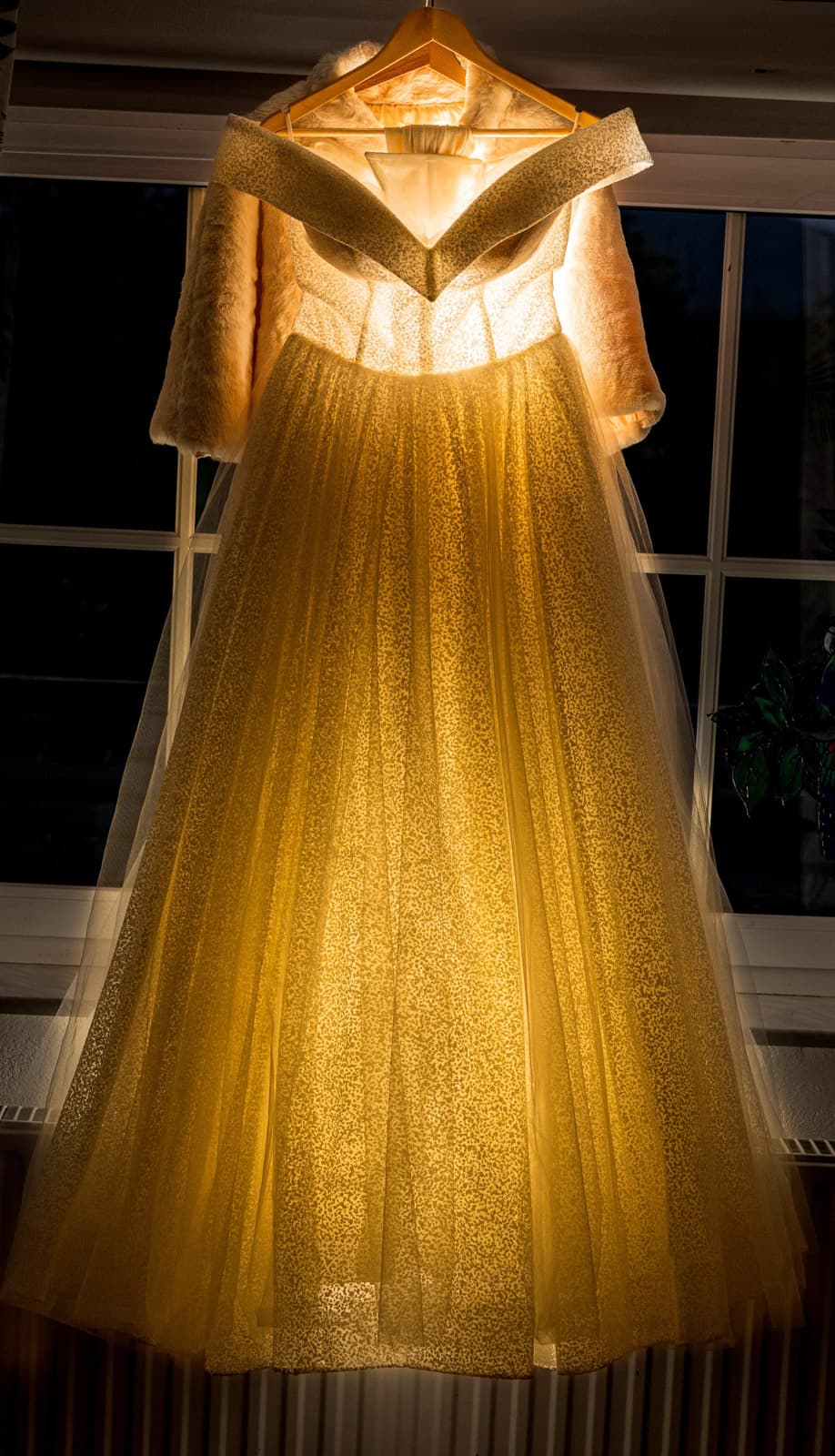 Glitzerndes Brautkleid auf goldenem Bügel vor abendlichem Sprossenfenster, von hinten beleuchtet