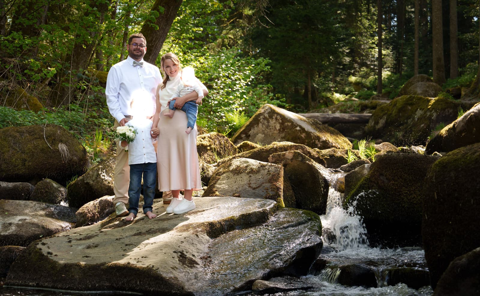 Familie mit Baby steht auf Felsblock im Waldbach, weiches Pastell-Sonnenlicht