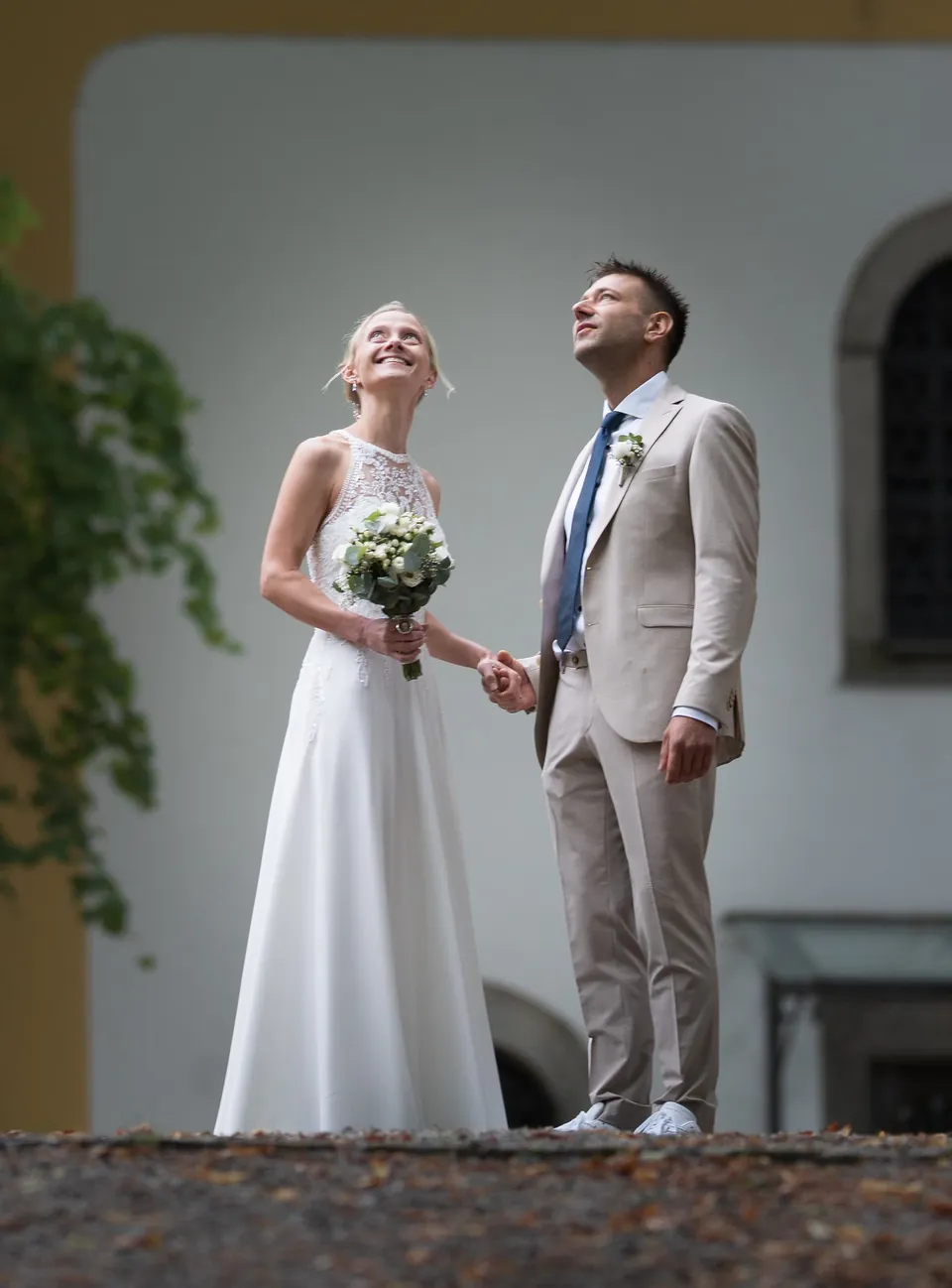 Brautpaar im weißen Kleid und hellen Anzug vor einem Gebäude.