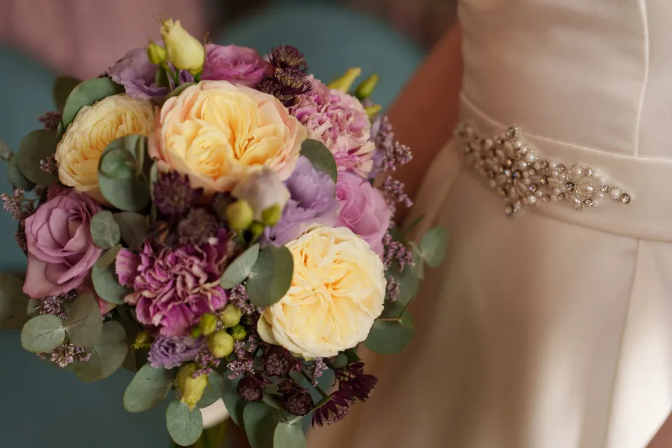 Brautstrauß mit gelben und violetten Blüten vor einem Brautkleid.